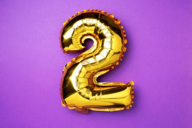 Creative layout. Golden foil balloon number and digit two 2. Birthday greeting card. Anniversary concept. Top view. Copy space. Stylish gold numeral over purple background. Numerical digit