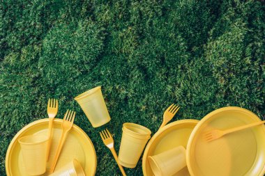 Environmental pollution. Disposable plastic tableware - plastic plates, forks, spoons, knives. Yellow plastic tableware on green grass, moss background. Top view. Copy space. Ban single use plastic.