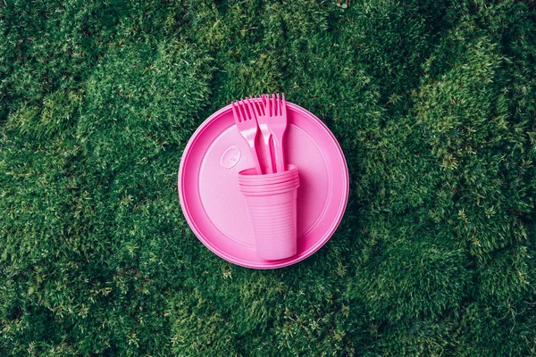 Pink plastic tableware on green grass, moss background. Top view. Copy space. Environmental pollution. Ban single use plastic. Disposable plastic tableware - plastic plates, forks, spoons, knives.