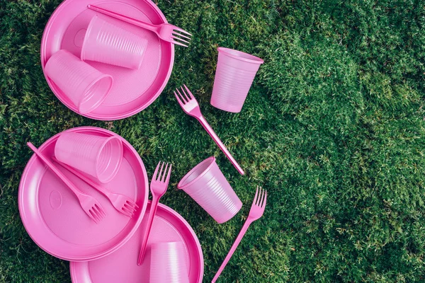 Pink plastic tableware on green grass, moss background. Top view. Copy space. Environmental pollution. Ban single use plastic. Disposable plastic tableware - plastic plates, forks, spoons, knives.