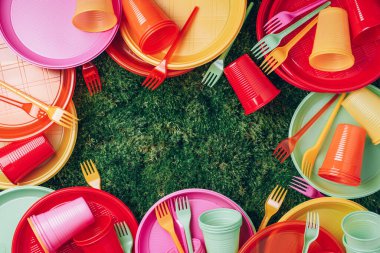 Colored plastic disposable tableware on green grass moss background. Top view. Copy space. Birthday picnic utensil. Recycling plastic and ecology. Disposable plastic tableware - plates, forks, spoons.