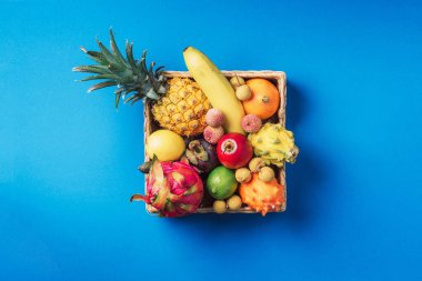 Mavi arka planda tropikal meyveler. Egzotik Tayland meyveleriyle dolu Rattan kutusu - ananas, pitahaya, kiwano, afrika boynuzlu kavun, tamarillo meyvesi, granadilla, feijoa, fıstık, yılan meyvesi, maracuya