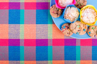 Fresh homemade muffins and cookies with color chocolate coated candies on a colorful checkered kitchen towel. Free space for your text