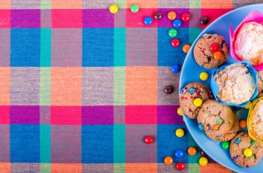 Fresh homemade muffins and cookies with color chocolate coated candies on a colorful checkered kitchen towel. Free space for your text