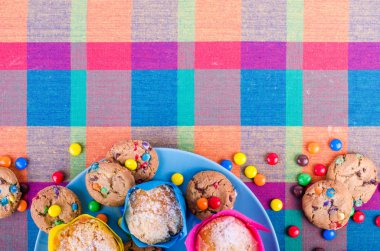 Fresh homemade muffins and cookies with color chocolate coated candies on a colorful checkered kitchen towel. Free space for your text