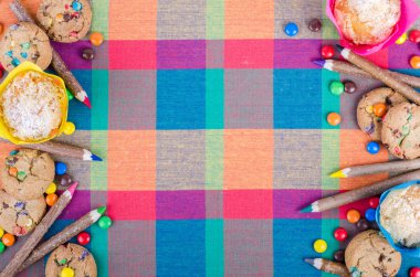 Fresh homemade muffins and cookies with color chocolate coated candies, colorful wooden pencils on a checkered kitchen towel. Free space for your text