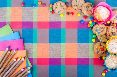 Fresh homemade muffins and cookies with color chocolate coated candies, colorful wooden pencils and notebooks, exercise books, cup of tea on a checkered kitchen towel. Free space for your text