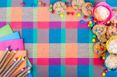 Fresh homemade muffins and cookies with color chocolate coated candies, colorful wooden pencils and notebooks, exercise books, cup of tea on a checkered kitchen towel. Free space for your text clipart