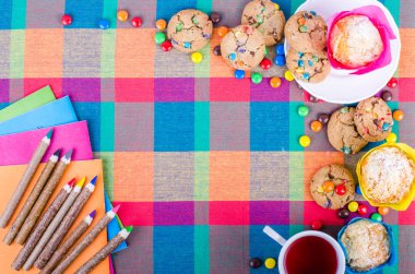 Fresh homemade muffins and cookies with color chocolate coated candies, colorful wooden pencils and notebooks, exercise books, cup of tea on a checkered kitchen towel. Free space for your text