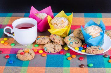 Fresh homemade muffins and cookies with color chocolate coated candies, cup of tea on a colorful checkered kitchen towel. Free space for your text