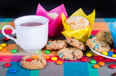 Fresh homemade muffins and cookies with color chocolate coated candies, cup of tea on a colorful checkered kitchen towel. Free space for your text