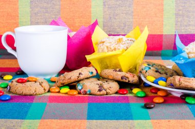 Fresh homemade muffins and cookies with color chocolate coated candies, cup of tea on a colorful checkered kitchen towel. Free space for your text