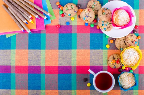 Frische hausgemachte Muffins und Kekse mit Schokoladenüberzug, bunte Holzstifte und Notizbücher, Schulhefte, eine Tasse Tee auf einem karierten Küchentuch. Freiraum für Ihren Text — Stockfoto