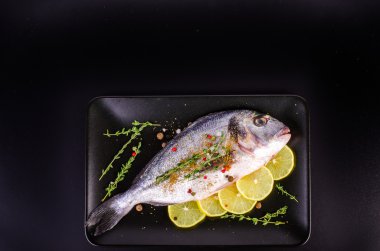 Fresh raw gilthead fishes with lemon, herbs, salt on plate, black background. Healthy food concept. Food frame
