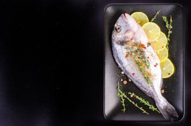 Fresh raw gilthead fishes with lemon, herbs, salt on plate, black background. Healthy food concept. Food frame
