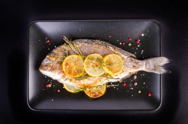 Roasted gilthead fishes with lemon, herbs, salt on black background. Healthy food concept. Food frame. Free space for your text.