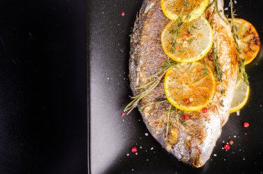 Roasted gilthead fishes with lemon, herbs, salt on black background. Healthy food concept. Food frame. Free space for your text.