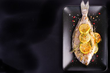 Roasted gilthead fishes with lemon, herbs, salt on black background. Healthy food concept. Food frame. Free space for your text.