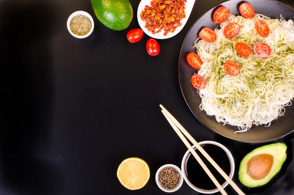 Tagliatelle di riso cinese con pesto, pomodorini ciliegia, avocado, salsa di soia, limone e spezie su sfondo nero. Spazio libero per il testo . — Foto Stock