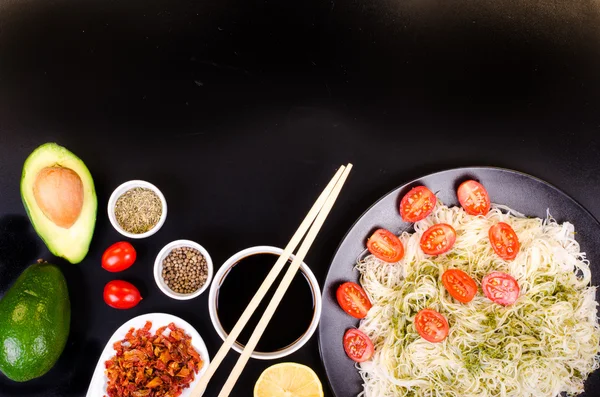 Tagliatelle di riso cinese con pesto, pomodorini ciliegia, avocado, salsa di soia, limone e spezie su sfondo nero. Spazio libero per il testo . — Foto Stock