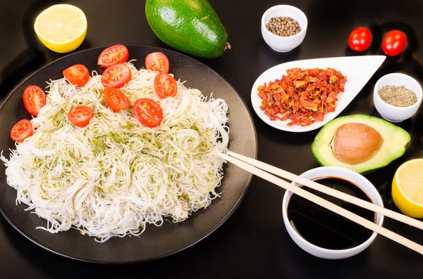 Tagliatelle di riso cinese con pesto, pomodorini ciliegia, avocado, salsa di soia, limone e spezie su sfondo nero . — Foto Stock