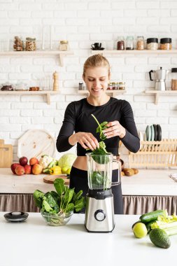 Sağlıklı beslenme, diyet konsepti. Genç, sarışın, gülümseyen kadın ev mutfağında yeşil smoothie yapıyor.