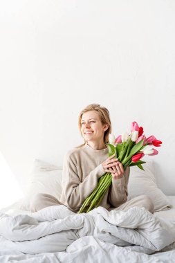 Mutlu kadın pijamalarıyla yatakta oturuyor, çiçeklerin tadını çıkarıyor.