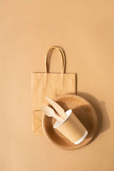 Brown craft disposable bag and tableware cup, plate, and wooden fork, knife on brown background flat lay top view