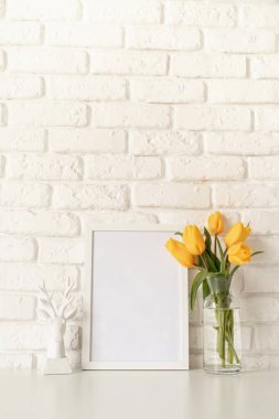 Cam bir vazoda sarı lale buketi, beyaz bir mum ve beyaz tuğla bir arka planda boş bir fotoğraf çerçevesi. Tasarımı düzenle