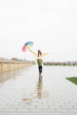 Sarı yağmurluklu güzel esmer kadın yağmurda gökkuşağı şemsiyesi tutuyor.