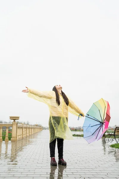 美しいですブルネット女性で黄色レインコート保持虹の傘アウトで雨 — ストック写真