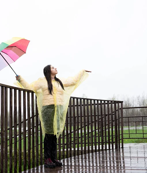 Beautiful smiling brunette woman in yellow raincoat holding rainbow umbrella out in the rain, catching raindrops with her hand