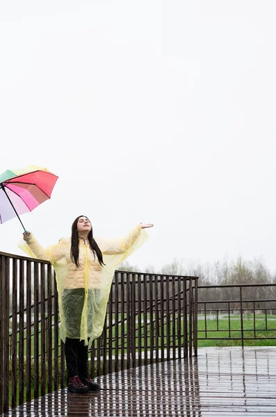 Beautiful smiling brunette woman in yellow raincoat holding rainbow umbrella out in the rain, catching raindrops with her hand