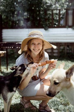 Yazlık şapkalı genç bir kadın arka bahçede et pişiriyor, köpeğiyle oturuyor, evcil hayvanına atıştırmalık veriyor.