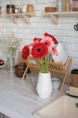 Beyaz tuğla duvar ve beyaz ahşap raflarla dolu taşralı bir mutfak. Beyaz vazoda taze Gerbera çiçekleri