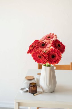 Beyaz vazodaki kırmızı ve pembe gerbera papatyaları. Mumlar ve fincanlarla birlikte, minimal tarzda.