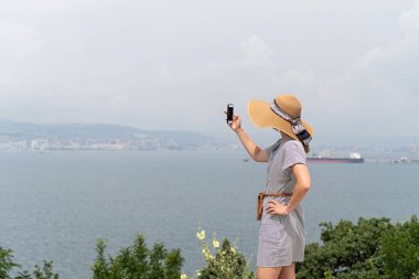 Yazlık elbiseli ve şapkalı genç bir kadının cep telefonu kullanarak şehir manzarasının fotoğrafını çekmesi. Özgürlük kavramı. Seyahat kavramı