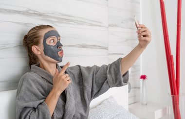 Kadın sağlığı. Spa ve sağlık. Yüz maskesi takan kadın yatağında mobil cihaz kullanarak rahatlıyor.