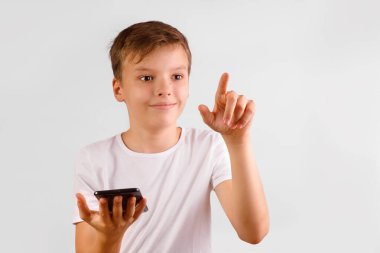Beyaz tişörtlü genç bir çocuk bir elinde koyu renk bir akıllı telefon tutuyor ve diğerinin işaret parmağıyla öne doğru işaret ediyor, hafifçe gülümsüyor, beyaz bir arka planda sanal ile etkileşim kurduğunu öne sürüyor..