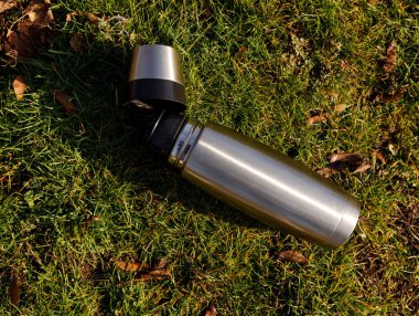 Kapağı çıkarılmış gümüş süpürge şişesi, dağınık yapraklarla yeşil çimlerin üzerine yerleştirilmiş. Güneşli sonbahar günlerinde kamp, yürüyüş ve açık hava kırılması için idealdir..