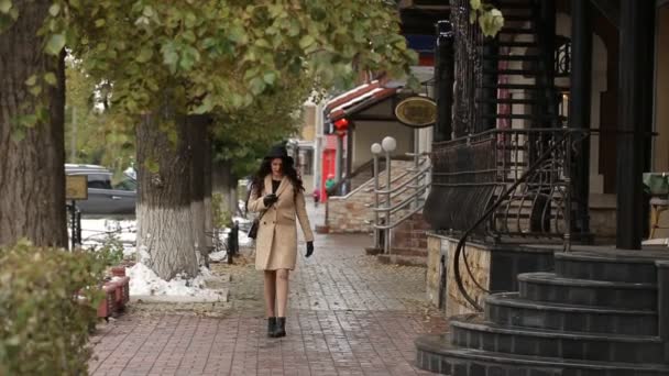 Девушка в бежевом пальто ходит и читает сообщение — стоковое видео