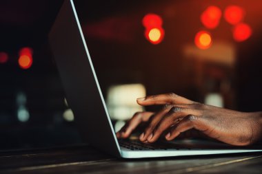 Siyah adam elleri klavyede yazarak. Dizüstü bilgisayarla çalışan kişi. Arka plan olarak güzel ışıklar. Tonda