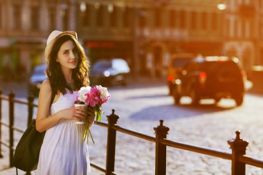 Young beautiful lady walking on street, holding paper cup with coffee and bouquet of peonies. Model wearing stylish clothes. City lifestyle. Copy space for text. Toned