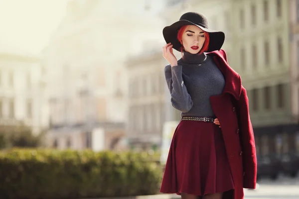 Portret młodej pięknej modnej kobiety pozowanie na ulicy. Model ubrany w stylowy czarny kapelusz z szerokim rondem, czerwony płaszcz. Dziewczyna patrzy w dół. Koncepcja mody kobiet. Kopiuj przestrzeń. Stonowanych — Zdjęcie stockowe