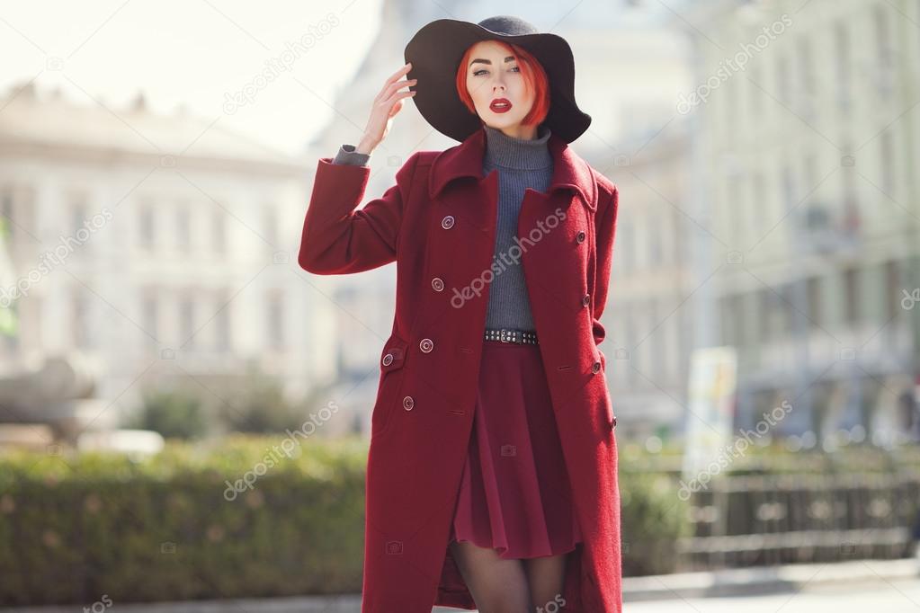 Young beautiful fashionable woman posing on the street. Model wearing stylish black wide-brimmed hat, red coat. Girl looking at camera. Female fashion concept. Toned