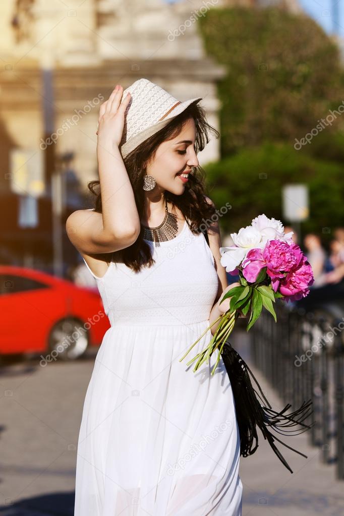 Outdoor portrait of young beautiful happy smiling lady walking on the street. Model wearing stylish white clothes, accessories. Girl looking down and aside. City lifestyle. Sunny day. Waist up.