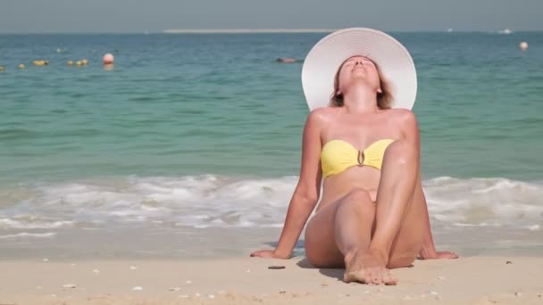 Portrait jeune femme souriante assise sur la plage en maillot de bain jaune et chapeau blanc — Video