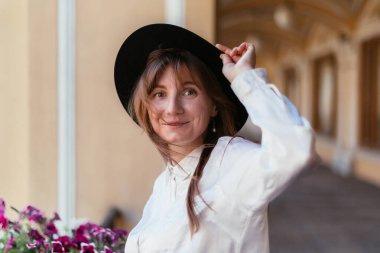 Beyaz kız elinde şapkayla gülümseyip kameraya bakıyor..