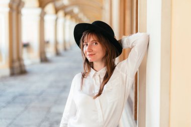 Şapkalı, gülümseyen ve binaya yaslanan beyaz bir kız..