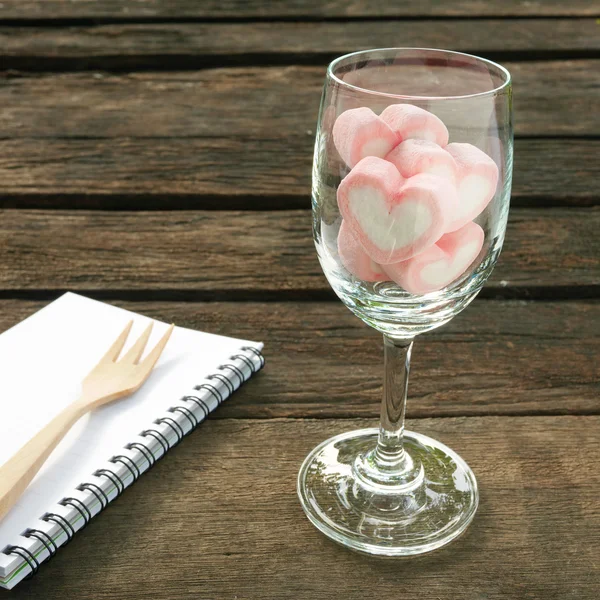 Il bel cuore rosa marshmallow in bicchiere di vino e libro di note bianche con forchetta in legno — Foto Stock